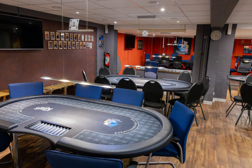Zwei Pokertische unter Deckenleuchten, an der Wand gerahmte Turnierfotos