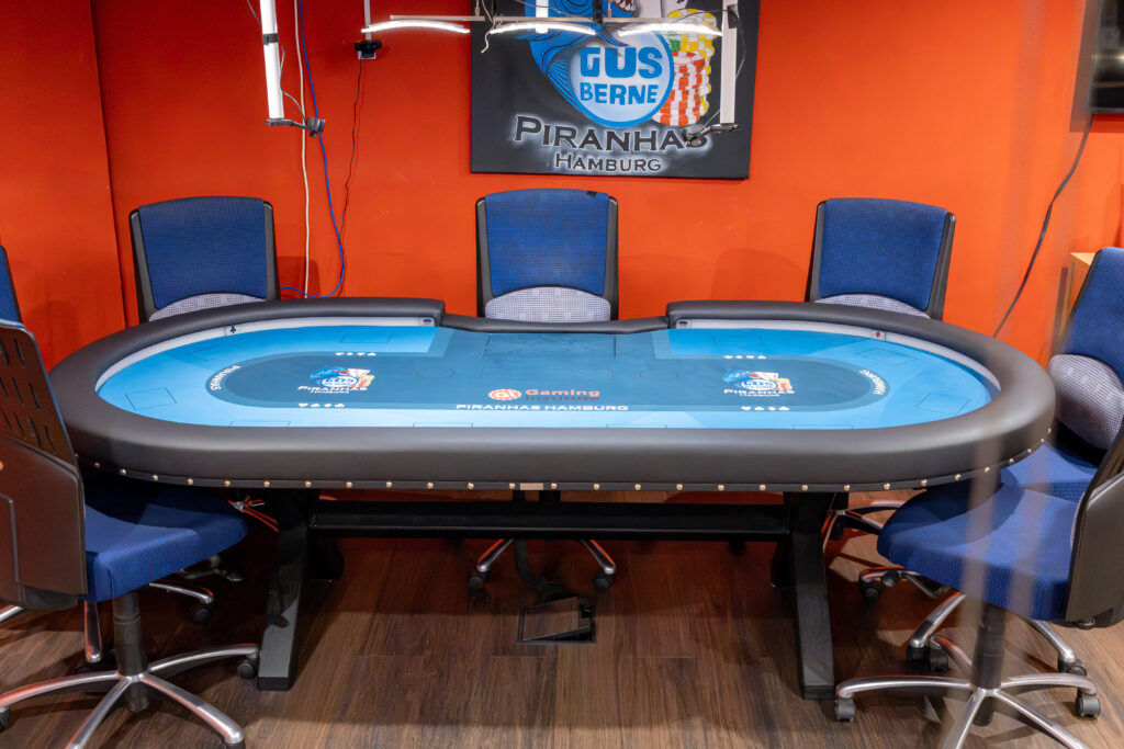 Pokertisch mit blauem Filz und aufgedrucktem Vereinslogo, dahinter an der orangefarbenen Wand das Wappen der Piranhas Hamburg